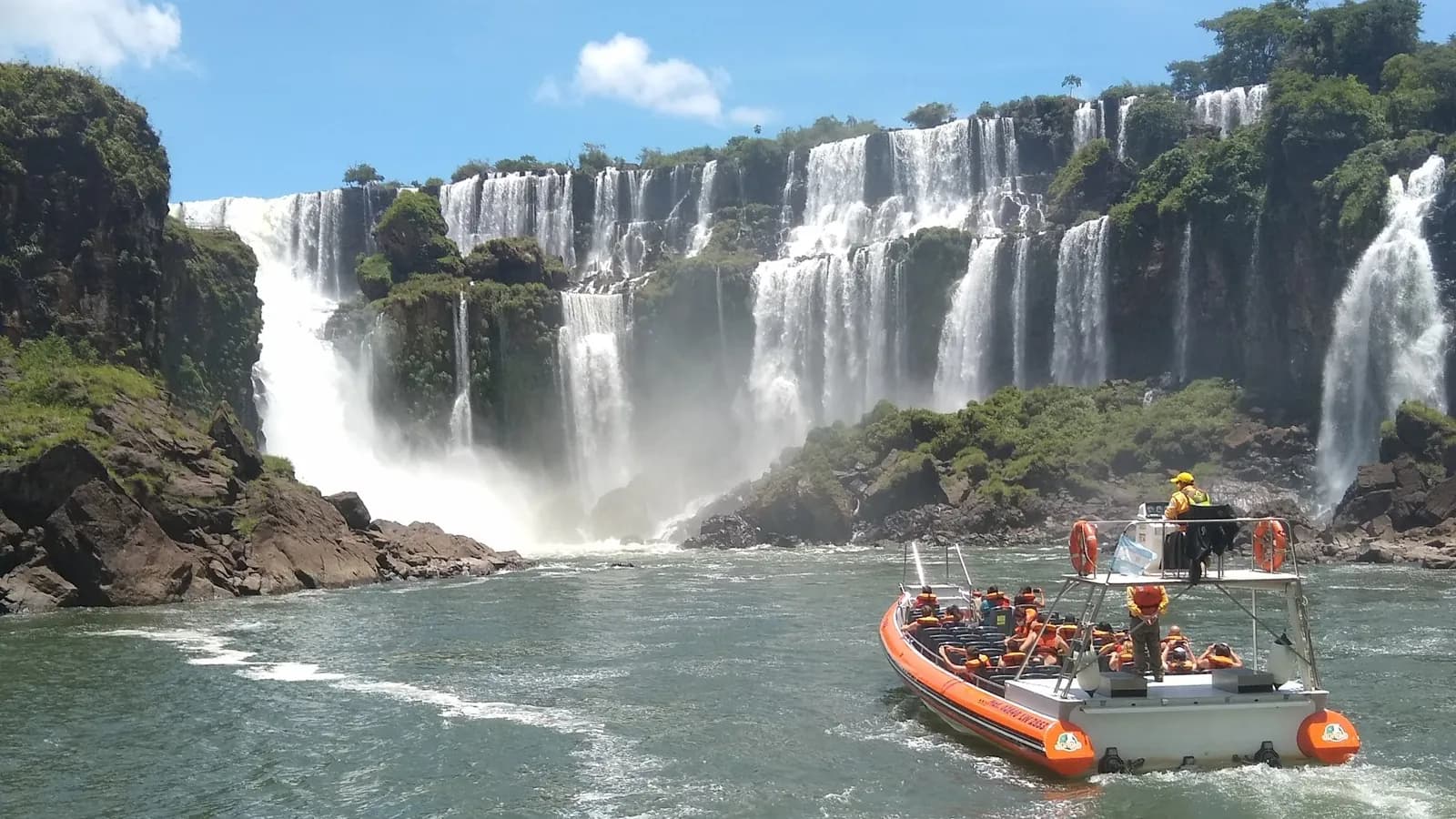 Orange Gran Aventura speedboat crossing the Iguazú River canyon towards a wide wall of waterfalls under a blue sky, Argentine side